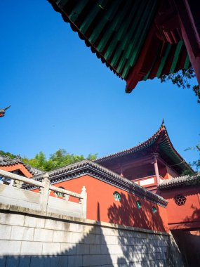 Geleneksel bir Çin binasının açık mavi gökyüzüne karşı karmaşık çatı yapısına, saçaklarına ve kırmızı duvarlarına bakıyorum..