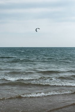 Ağır gri gökyüzünün altında dalgalı sularda gezinen tek bir uçurtma sörfçüsünü yakalayan dikey bir çekim. Havadaki uçurtmayı aydınlatıyor..