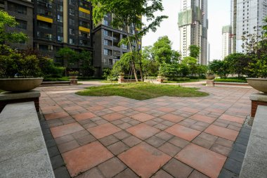 Ağaçlar ve modern yerleşim yerleriyle çevrili, kırmızı ve gri geometrik fayanslarla döşenmiş geniş bir kamu avlusunun geniş açılı görüntüsü.