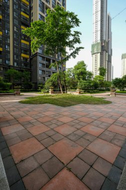 Merkezi bir ağaç ve ekicilerin yer aldığı fayanslı bir açık meydanın dikey perspektifi, mavi gökyüzüne karşı yüksek apartman gökdelenlerine doğru bakıyor..