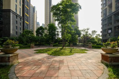 Merkezi ağaçla çevrili dairesel asfalt bir avlunun göz hizasında, bakımlı bahçeler ve modern apartman blokları ile çevrili..