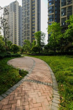 Yuvarlak kırmızı tuğlalı bir geçitin dikey görüntüsü yeşil bir çimenliğin içinden geçip modern yüksek katlı apartmanlara doğru gidiyor..