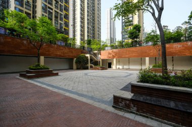 Kırmızı tuğlalı istinat duvarlarıyla karakterize edilen modern bir halk meydanı, ağaçlı yerleşik ekiciler, ve binalarla çevrili geometrik asfalt zemin..