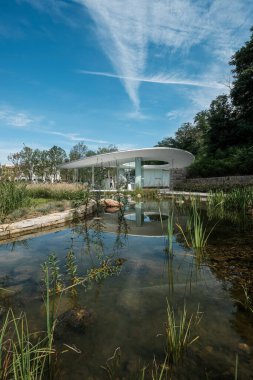 Sazlıklar ve yansımalarla dolu sakin bir sulak göletin dikey manzara fotoğrafında, arka planda mavi bir gökyüzünün altında modern kıvrımlı beyaz bir pavyon var..