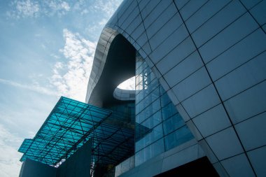 Kıvrımlı cam perde duvarı, metal bir tepe örtüsü ve mavi gökyüzüne karşı dairesel bir açıklığı olan modern bir binanın düşük açılı mimari detayları..