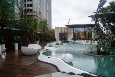 İçinde sığ bir havuz, ahşap bir güvertede beyaz mobilyalar ve büyük bir metal heykel bulunan lüks bir açık hava eğlence alanı..