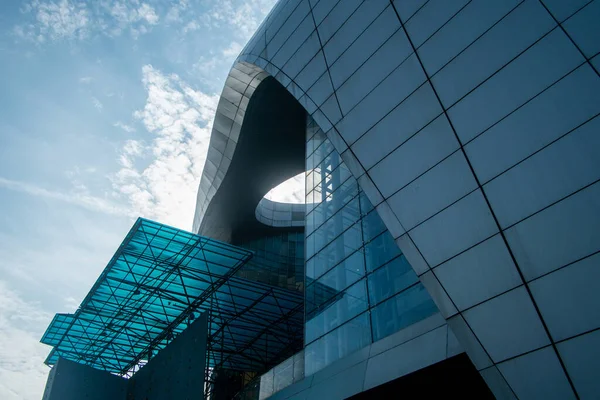 Kıvrımlı cam perde duvarı, metal bir tepe örtüsü ve mavi gökyüzüne karşı dairesel bir açıklığı olan modern bir binanın düşük açılı mimari detayları..