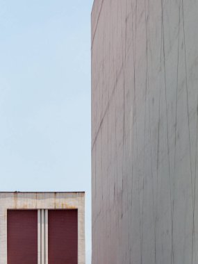 Dikey mimari detay, gri beton bir duvar ile açık mavi gökyüzüne karşı kırmızı ve beyaz bir binanın geometrik bir bölümü arasındaki katı zıtlığı gösteriyor..