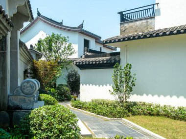 Beyaz duvarların, geleneksel çatı çizgilerinin, taş elementlerin ve yemyeşil bitkilerin bulunduğu Çin avlusundaki sessiz bir köşenin yatay görüntüsü.