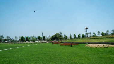 Manikürlü yeşil bir çim parkının geniş bir manzara görüntüsü, kırmızı çiçek yataklarıyla birlikte, engin bir mavi gökyüzüne karşı,.