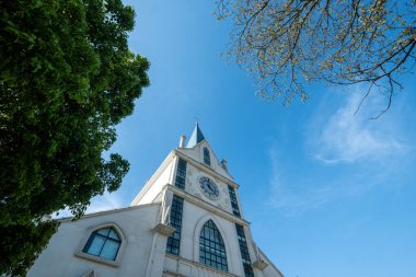 Düşük açılı perspektif beyaz bir kilise kulesi ve çan kulesine bakıyor, açık mavi gökyüzüne karşı yeşil ağaç dalları tarafından çerçevelenmiş..