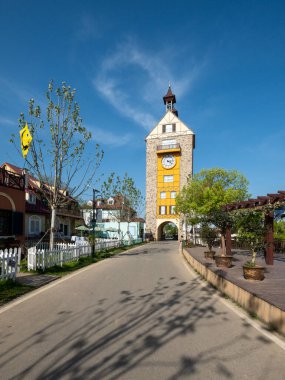 İnce bulutlu mavi gökyüzünün altındaki büyük sarı saat kulesine doğru uzanan asfalt bir yolun dikey bakış açısı.