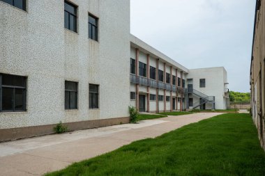 Beyaz ya da açık gri beton bir binanın dış görünüşü, muhtemelen bir okul ya da ofis, sıralı pencereler ve iyi korunmuş yeşil çimenler.