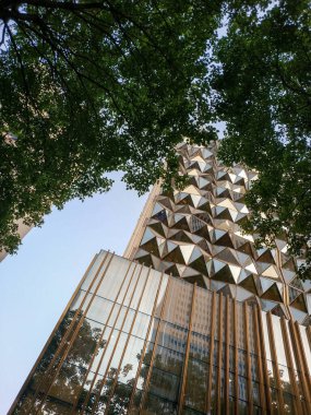 Modern bir gökdelenin tepesine bakıyorum. Karmaşık geometrik kaplama ve yansıtıcı camlar açık mavi gökyüzüne karşı..