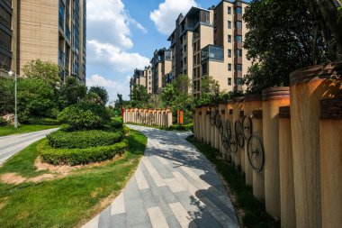 Bir apartman bahçesinde bisiklet temalı sanat eserlerinin yer aldığı dekoratif bir duvarın yanında uzanan bir yaya yolunun yatay görüntüsü..