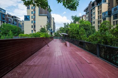 Ağaçlar ve konut binalarıyla çevrili bir apartman parkında uzanan kırmızı tahta bir köprü manzarası..
