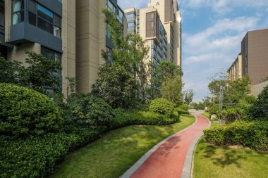 Modern bir apartman parkında manikürlü çimlerin ve ağaçların arasından geçen kırmızı bir yürüyüş yolunun manzarası..