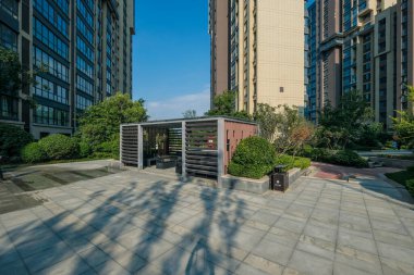 Modern, minimalist ahşap bir çardağın manzarası. Parlak, taş döşeli bir plazanın içinde bir apartman kompleksi..