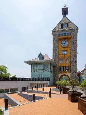 Avrupa tarzı geleneksel bir saat kulesinin dikey görüntüsü, ön planda manzaralı bir avlunun yer aldığı temalı bir turizm parkı içerisindedir..