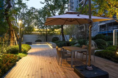 Modern mobilyaları olan, sakin bir akşam bahçesinin arka planına sıcak bir şekilde aydınlatılmış davetkar bir oturma alanı. Görüntü huzur, yaşam tarzı, lüks ve huzurlu bir atmosfer sağlıyor. Sığ alan derinliğine sahip yüksek kaliteli gerçek fotoğrafçılık.