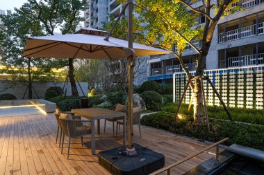 Bahçe manzaralı bir bahçede sıcak akşam ışıklarıyla aydınlatılmış bir şemsiyenin altında huzurlu bir dinlenme odası düzenlemesi. Görüntü rahatlamayı, konforu, modern yaşamı ve dingin ambiyansı aktarır. Sığ alan derinliğine sahip yüksek kaliteli gerçek fotoğrafçılık