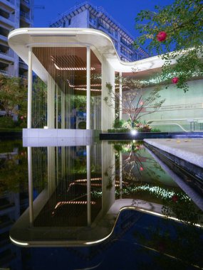 Geceleri sakin ve yansıtıcı bir havuza karşı parlayan modern bir pavyonun yer aldığı geniş bir bahçe manzarası. Görüntü huzur, mimari lüks, farkındalık ve sakin bir akşam atmosferi sağlıyor. Sığ alan derinliğine sahip yüksek kaliteli gerçek fotoğrafçılık