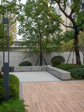 Ahşap bir güverte, taş banklar ve manikürlü ağaçların yer aldığı çağdaş bir şehir avlusu sessiz bir geri çekilme yaratıyor. Görüntü lüks yaşam, açık hava konforu ve uzamsal uyum sağlıyor. Sığ alan derinliğine sahip yüksek kaliteli gerçek fotoğrafçılık.