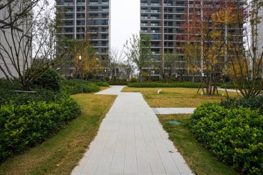 Modern bir yerleşim bölgesinde çalılarla çevrili yeşil bir çimenlikten geçen düz taşlı patika. Görüntü, huzurlu bir mahalle ve huzurlu bir ortam sağlıyor. Doğal ışıklandırmalı yüksek kaliteli gerçek fotoğrafçılık.