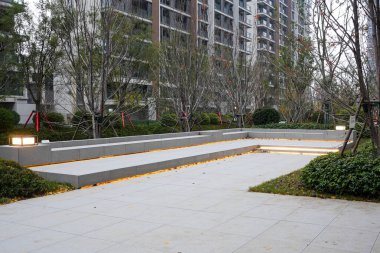 Alacakaranlıkta sakin bir topluluk ortamında sıcak LED şeritlerle aydınlatılan basamakları içeren asfalt taşlı plazanın geniş manzarası. Görüntü, kentsel sükunet ve modern peyzaj tasarımı sağlıyor. Doğal ışıklandırmalı yüksek kaliteli gerçek fotoğrafçılık.