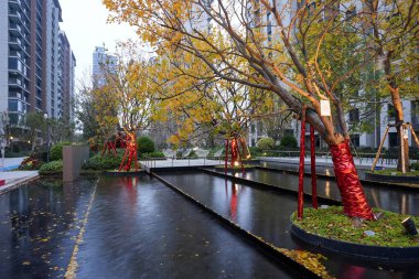 Modern apartman binalarını yansıtan sakin manzara havuzu ve alacakaranlıkta sakin bir halk avlusunda sonbahar sarı yaprakları. Görüntü, kentsel vahayı ve barışçıl atmosferi taşır. Doğal ışıklandırmalı yüksek kaliteli gerçek fotoğrafçılık.