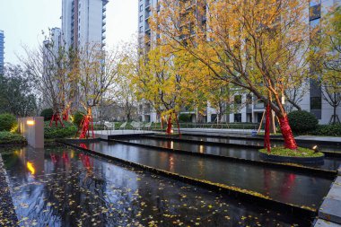 Sonbahar ağaçlarıyla çevrili geniş bir manzara havuzu. Alacakaranlık sırasında modern bir yerleşim yerinde sarı yapraklar var. Resim, huzurlu bir toplum ve zarif bir yaşam ortamı sunuyor. Doğal ışıklandırmalı yüksek kaliteli gerçek fotoğrafçılık.
