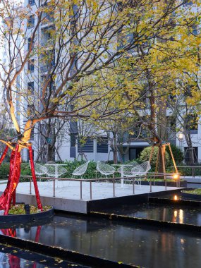 Aydınlatılmış açık hava terası, dingin bir yansıtma havuzunun yanında beyaz tel sandalyeler ve alacakaranlıkta sonbahar ağaçları. Görüntü rahat bir mahalle ve zarif bir toplum tasarımı sunuyor. Doğal ışıklandırmalı yüksek kaliteli gerçek fotoğrafçılık.