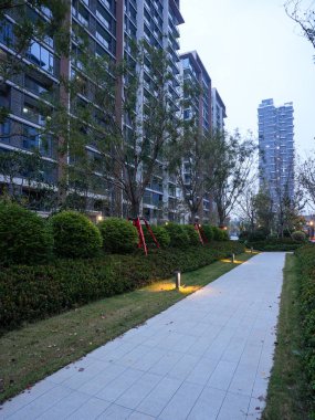 Yeşil bir çimenlik ve alacakaranlıkta yüksek binaların yanında parlayan zemin ışıkları olan düz asfalt bir yol. Görüntü, şehir sakinliğini ve modern yaşamı aktarıyor. Doğal ışıklandırmalı yüksek kaliteli gerçek fotoğrafçılık.