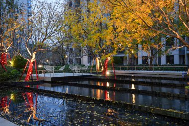 Mavi saat boyunca dingin bir manzara havuzuna yansıyan ılık ışıklarla aydınlatılan parlak sarı sonbahar yaprakları. Görüntü, ılık bir ortam ve mevsimsel güzelliği yansıtıyor. Doğal ışıklandırmalı yüksek kaliteli gerçek fotoğrafçılık.