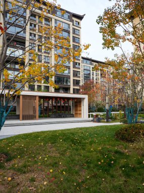 Huzurlu bir avluda, modern ahşap bir pavyon ve yeşil bir çimenlik üzerinde yükselen yüksek binalar. Görüntü, kentsel yaşam ve huzur dolu bir mahalleyi temsil ediyor. Doğal ışıklandırmalı yüksek kaliteli gerçek fotoğrafçılık.