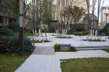 Huzurlu bir yerleşim bölgesinde yeni dikilmiş ağaçların etrafına inşa edilmiş dairesel beyaz bankların yer aldığı modern asfalt plaza. Görüntü, topluluk toplanmasını ve çağdaş manzara tasarımını iletir. Doğal ışıklandırmalı yüksek kaliteli gerçek fotoğrafçılık.