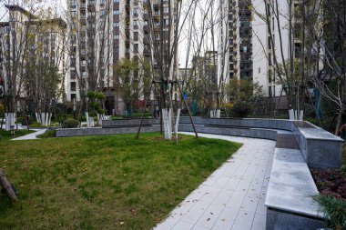 Modern bir halk avlusunda asfalt bir yolun yanındaki yeşil çimenliği çevreleyen açısal taş istinat duvarı. Görüntü, yapılandırılmış peyzaj tasarımı ve sakin şehir yaşamını taşır. Doğal ışıklandırmalı yüksek kaliteli gerçek fotoğrafçılık.