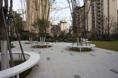 Asfaltlı bir avluda bir ağaç gövdesinin etrafına inşa edilmiş modern dairesel beyaz oturma düzeninin ayrıntılı görüntüsü. Görüntü çağdaş tasarım ve zarif açık hava mobilyalarını taşıyor. Doğal ışıklandırmalı yüksek kaliteli gerçek fotoğrafçılık.