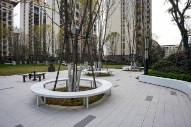 Geniş asfalt bir plazadaki bir ağacı çevreleyen modern dairesel beyaz bankın simetrik görüntüsü. Görüntü açık hava eğlencesi ve ahenkli bir toplum alanı sunuyor. Doğal ışıklandırmalı yüksek kaliteli gerçek fotoğrafçılık.