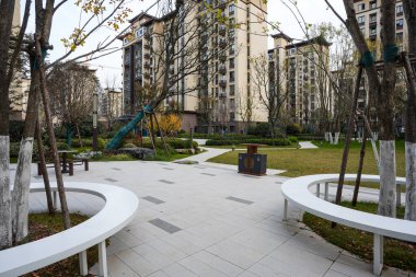 Asfaltlı bir plazadaki bir ağacın etrafındaki modern dairesel beyaz bankın arka planda bir oyun parkı görüntüsü. Görüntü, aktif bir mahalle ve aile dostu ortam sağlıyor. Doğal ışıklandırmalı yüksek kaliteli gerçek fotoğrafçılık.