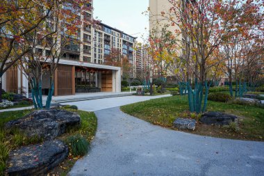 Bahçe yolunun manzarası, dekoratif kayalar ve modern halk köşkünü çevreleyen canlı sonbahar ağaçları. Görüntü huzurlu bir ortam ve uyumlu bir şehir hayatı sunuyor. Doğal ışıklandırmalı yüksek kaliteli gerçek fotoğrafçılık.