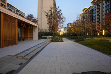 Geniş asfaltlı halk meydanı, alacakaranlıkta sonbahar ağaçlarıyla çevrili sıcacık, ahşap bir pavyona açılıyor. Görüntü huzurlu bir akşam ve lüks bir mahalle tasarımı sunuyor. Doğal ışıklandırmalı yüksek kaliteli gerçek fotoğrafçılık.