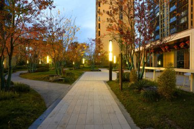 Taş kaldırımlı kaldırım yolu çakıl yolu ile kesişiyor, alacakaranlıkta halka açık bir bahçede sıcak ışıklandırılmış sokak lambaları ve sonbahar yapraklarıyla çevrili. Görüntü, huzurlu bir mahalle ve güvenli bir ortam sağlıyor. Doğal ışıklandırmalı yüksek kaliteli gerçek fotoğrafçılık