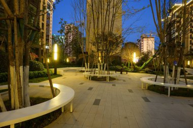 Asfalt bir plazadaki ağaçların etrafına inşa edilmiş beyaz dairesel banklar, derin mavi alacakaranlık gökyüzüne karşı sıcak elektrik direkleriyle aydınlatılıyor. Görüntü, topluluk toplanması ve çağdaş peyzaj mimarisini taşır. Doğal li ile yüksek kaliteli gerçek fotoğrafçılık