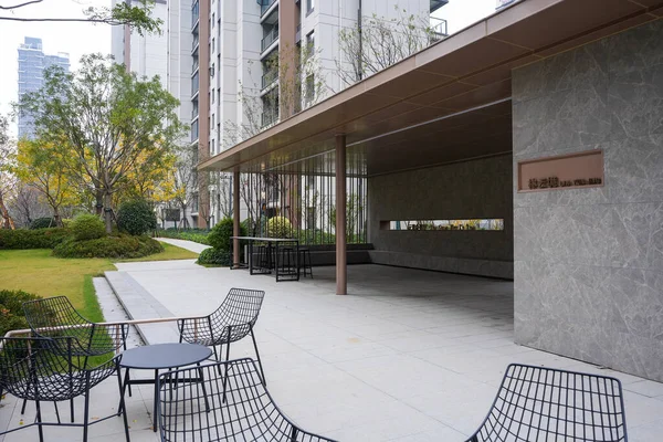 Modern bir yerleşim bölgesinde aynı metal masalar ve sandalyelerle geniş bir açık hava oturma alanı. Görüntü açık alan ve mahalle uyumu taşır. Doğal ışıklandırmalı yüksek kaliteli gerçek fotoğrafçılık.