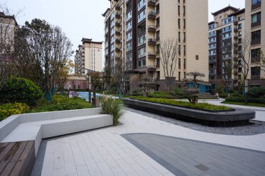 Kavisli taş koltuklu çağdaş asfaltlı avlu ve bakımlı bir yerleşim bölgesinde ekiciler yetiştirildi. Görüntü, yapılandırılmış manzara ve modern komşuluk yaşamını aktarıyor. Doğal ışıklandırmalı yüksek kaliteli gerçek fotoğrafçılık.