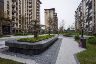 Modern bir avluda, dekoratif çakıl ve asfalt kaldırımlarla çevrili küçük ağaçlarla dolu uzun, karanlık bir taş ekicisi. Görüntü zarif bir açık hava tasarımı ve huzurlu bir toplum ortamı sunuyor. Natu ile yüksek kaliteli gerçek fotoğrafçılık