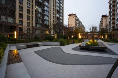 Alacakaranlıkta kavisli taş oturma alanlarıyla modern asfalt avluyu aydınlatan kalın ışık. Görüntü, kentsel sükunet ve zarif açık hava konforlarını iletiyor. Doğal ışıklandırmalı yüksek kaliteli gerçek fotoğrafçılık.