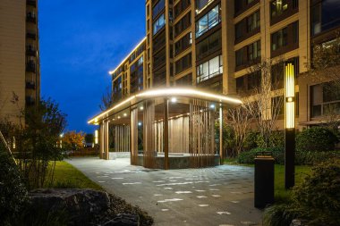 Çağdaş ahşap bir pavyonun ışıltılı kenarı, asfalt kaldırımın yanında ve derin mavi bir gökyüzünün altında uzun bir apartman dairesi. İmaj, kentsel zarafet ve lüks toplum konforlarını iletiyor. Doğal ışıklandırmalı yüksek kaliteli gerçek fotoğrafçılık.