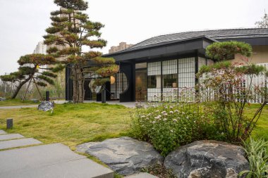 Çok güzel şekilli çam ağacı ve büyük dekoratif taşlar. Çağdaş bir binanın dışında, şebeke pencereli yeşil bir çimenlikte. Görüntü, doğunun zarafetini ve düşünceli yaşamını yansıtıyor. Doğal ışıklandırmalı yüksek kaliteli gerçek fotoğrafçılık.